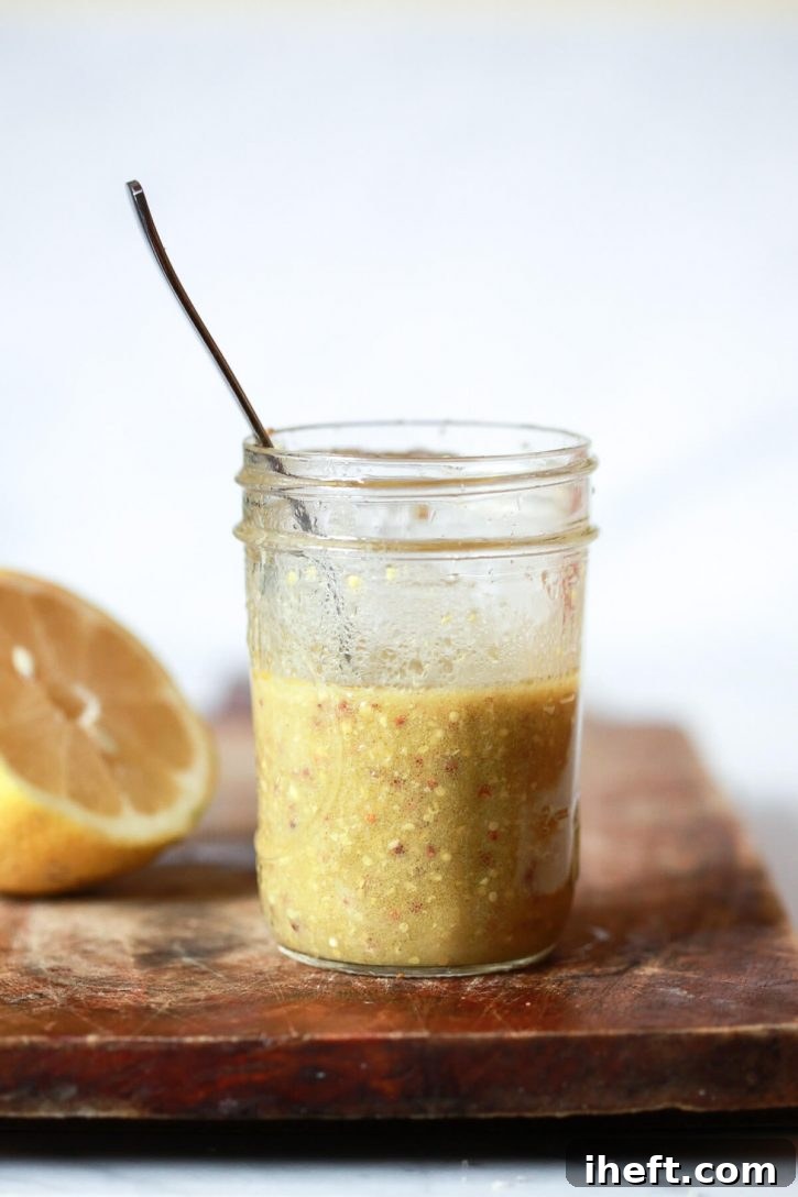 A small jar filled with a homemade whole seed mustard vinaigrette sits on a wooden cutting board with a lemon in the background. 