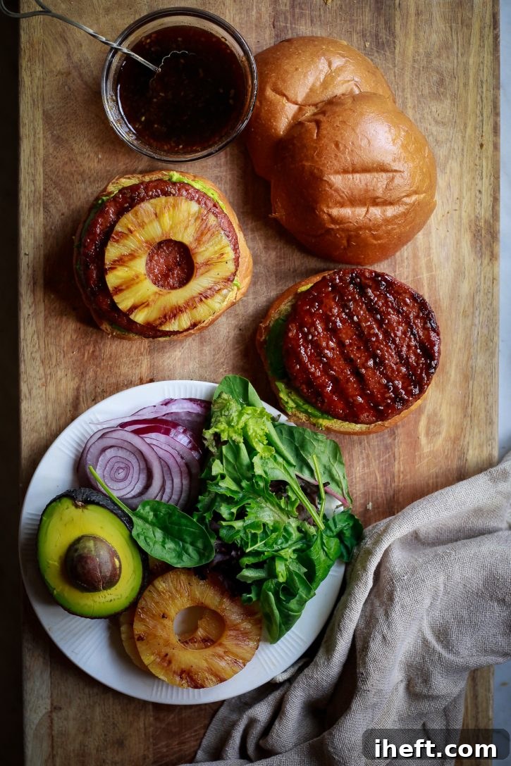 Pineapple teriyaki grilled veggie burgers are assembled on a wooden cutting board. Ingredients include grilled pineapple rings, greens, avocado, and red onion.