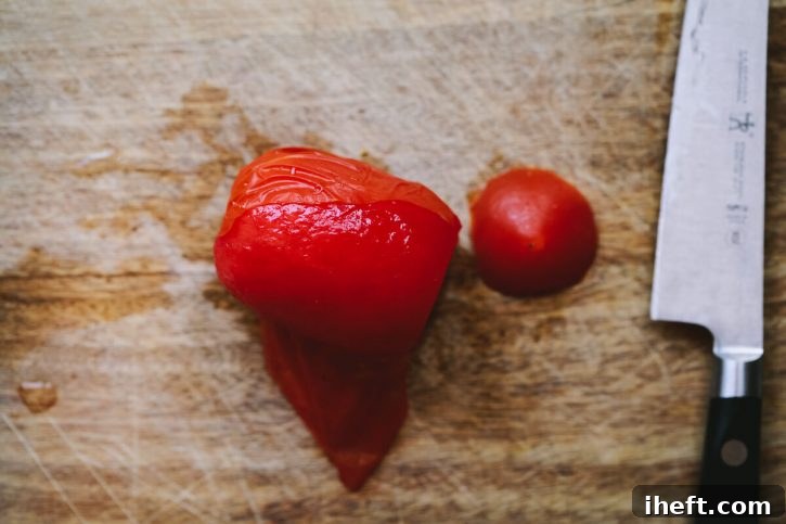 A Roma tomato that has been frozen and thawed is being peeled on a cutting board, illustrating the ease of skin removal after freezing.