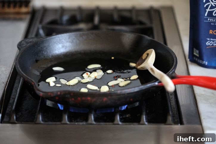 Thinly sliced garlic cloves and a pinch of vibrant red pepper flakes gently sauté in shimmering olive oil within a seasoned cast iron skillet, infusing the oil with their potent aromas and creating the flavorful base for the pasta dish.