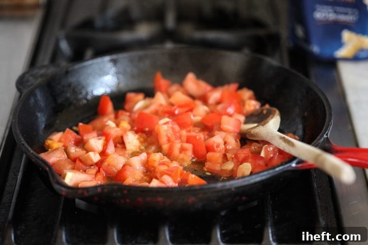 Freshly diced red tomatoes sizzle gently in a cast iron skillet, their juices starting to release and mix with the aromatic olive oil and garlic, creating a vibrant and savory base for the pasta sauce.
