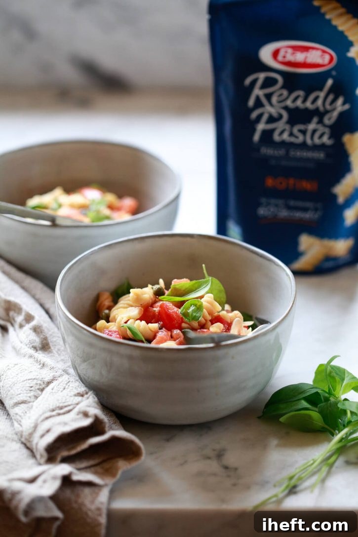 A generously sized bowl brimming with freshly prepared tomato basil pasta sits invitingly on a polished marble countertop. This vegetarian and vegan-friendly dish, crafted in minutes, features a harmonious blend of tender pasta, ripe tomatoes, fragrant basil, savory garlic, and zesty capers, showcasing a quick and healthy meal option.