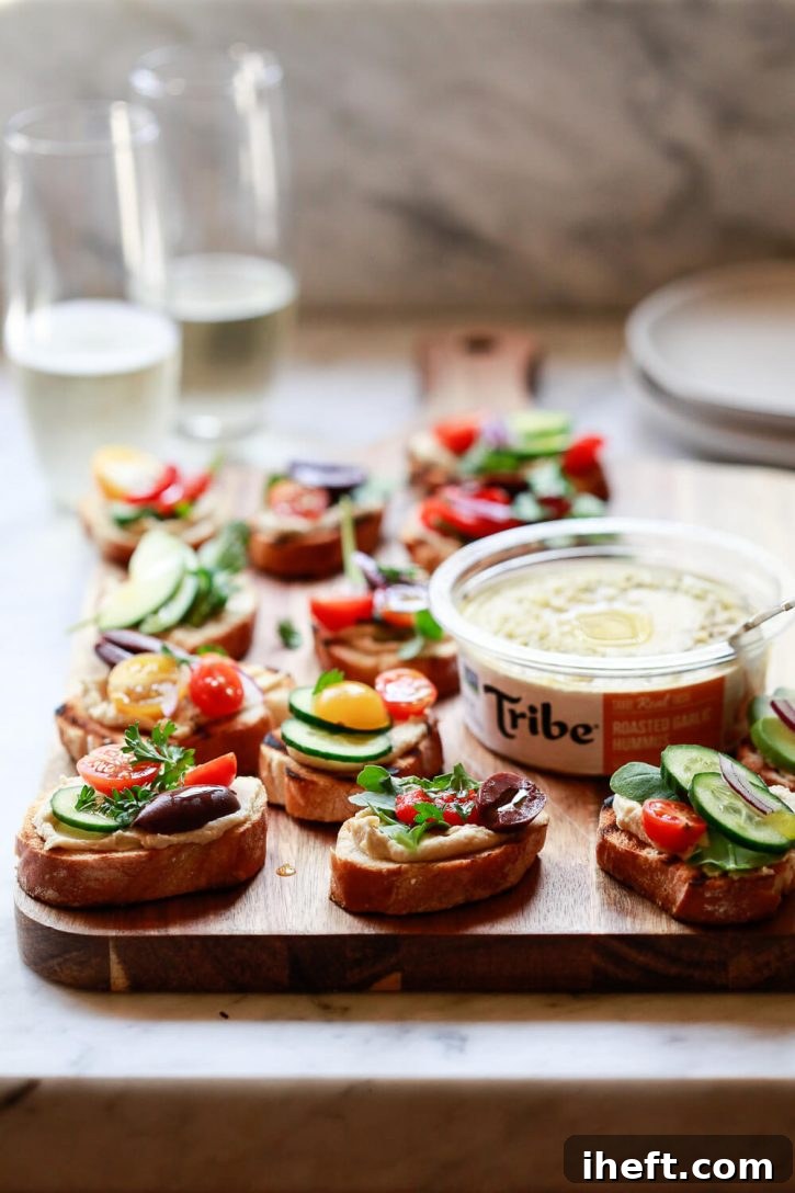 Crostini topped with hummus, olives, cucumber, and tomatoes sit on a cutting board. A beautiful vegan appetizer perfect for any occasion.