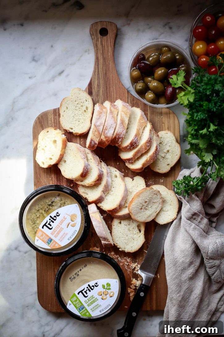 A French baguette is expertly sliced into uniform rounds, placed on a cutting board alongside two tubs of Tribe hummus, a bowl of olives, and cherry tomatoes, illustrating the initial steps of crostini preparation.