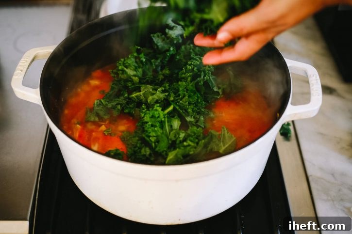 A hand gently drops fresh, vibrant kale into a pot of richly simmering Tuscan Ribollita, ready to wilt into the hearty vegetable and bean stew.