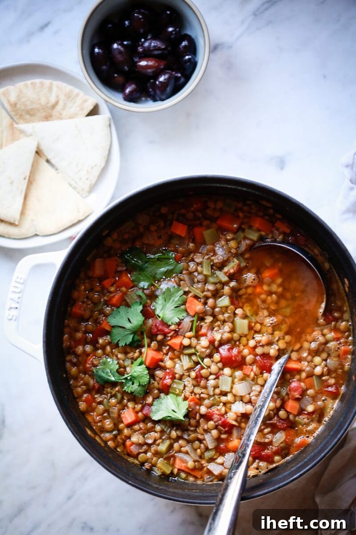 A white Staub pot filled with fakes/Greek lentil soup and a ladle. This is a healthy vegan lentil soup recipe. It sits next to pita bread and olives. 