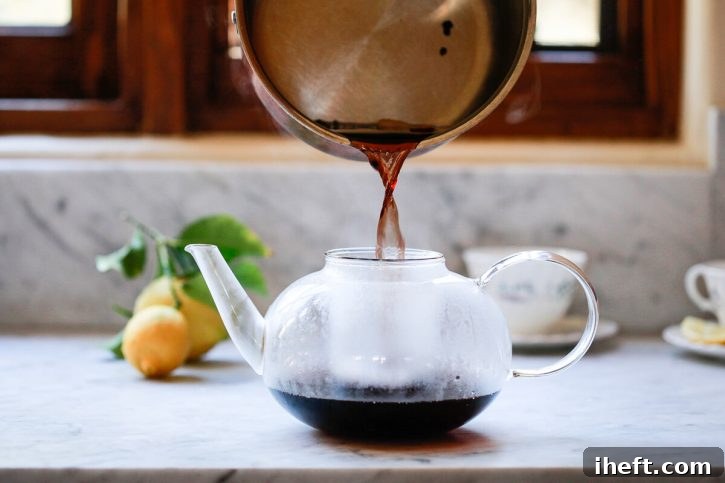 A saucepan of homemade elderberry tea is poured through a strainer in a teapot. 