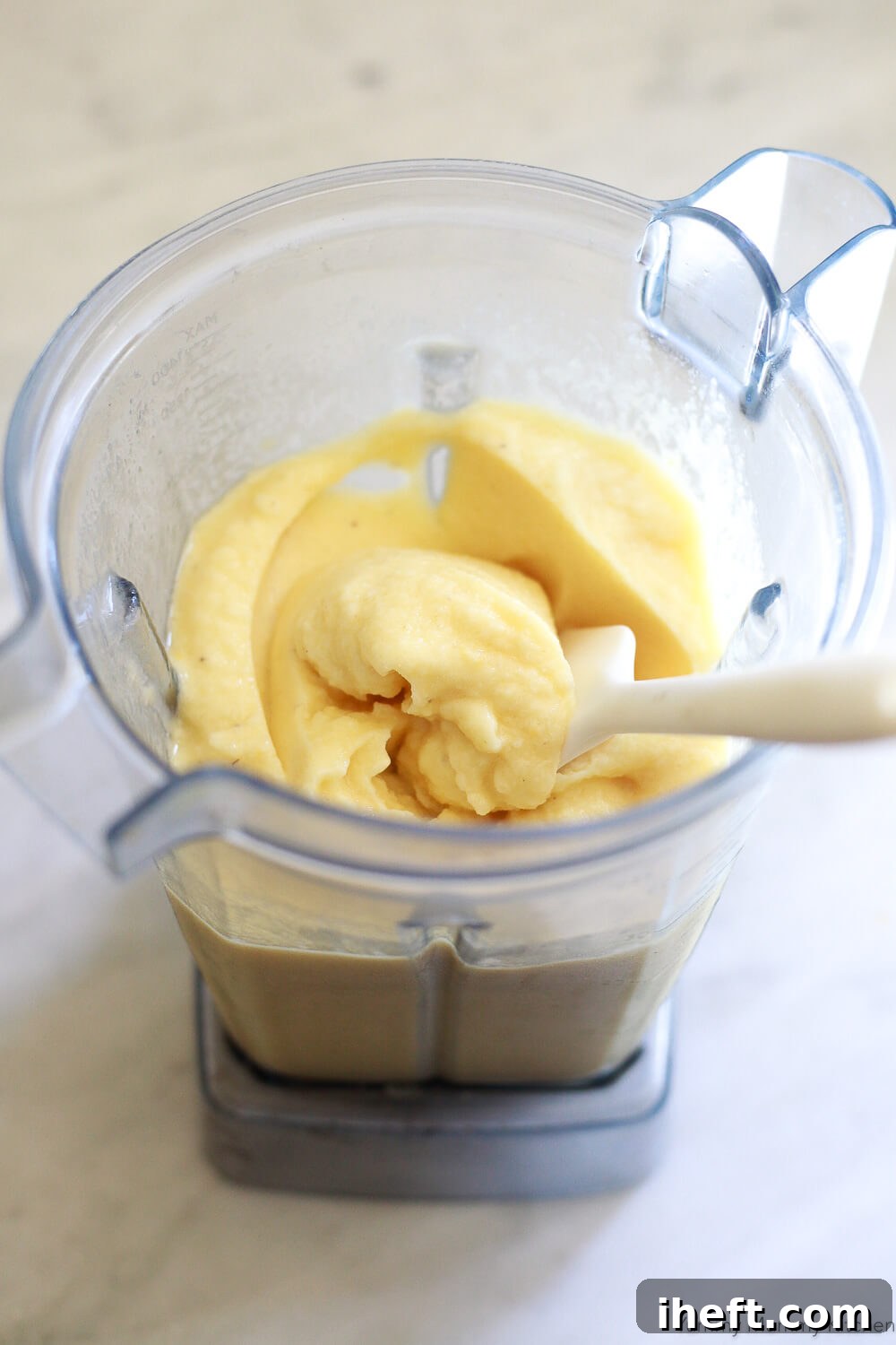 A very thick pineapple smoothie is scooped with a rubber spatula inside a Vitamix blender. 