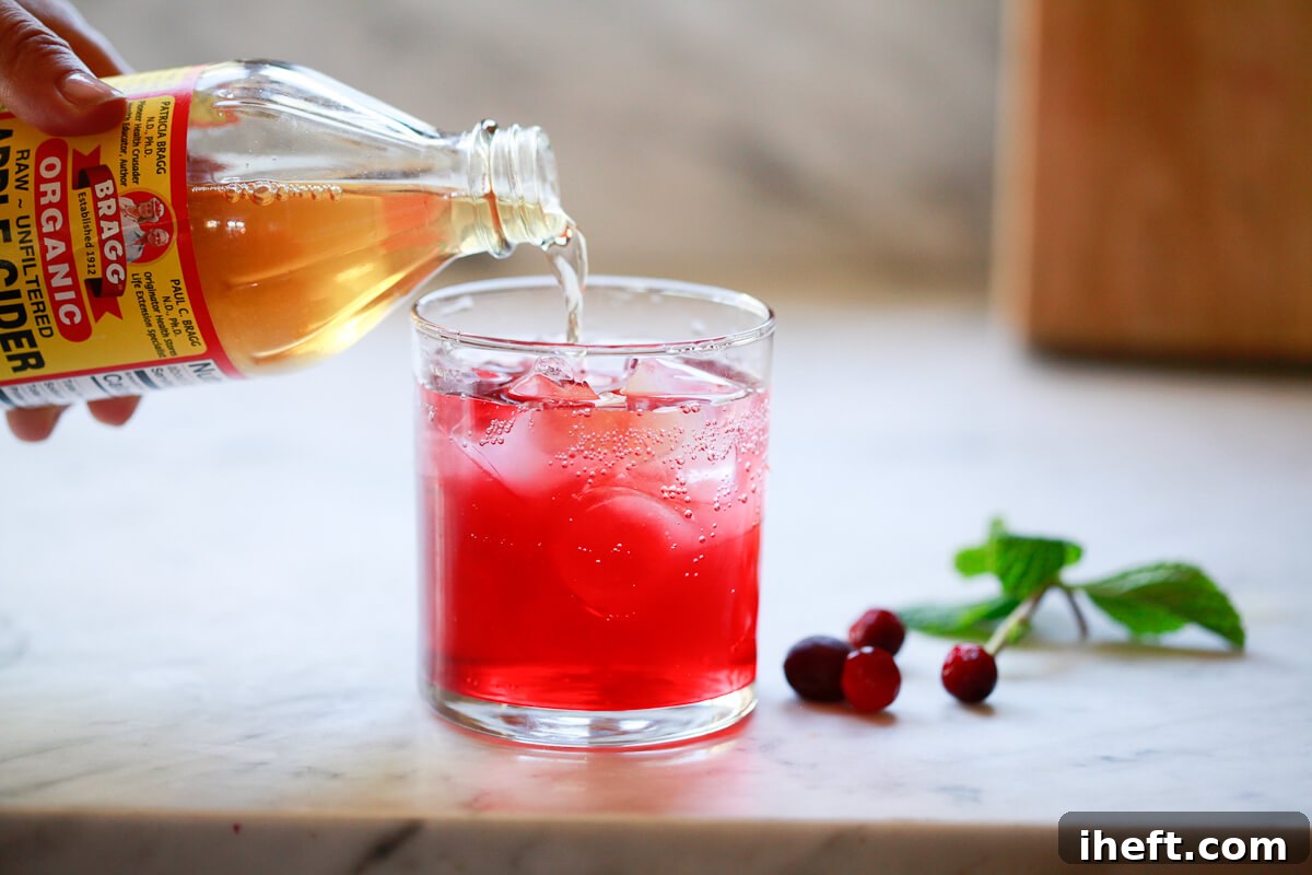 Bragg's apple cider vinegar is carefully poured into a glass already containing cranberry juice and water, over ice.