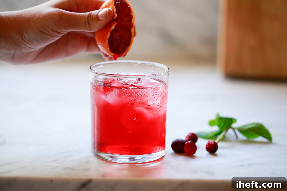 A hand gently squeezes half a blood orange into a cranberry detox drink, adding a fresh citrus note.