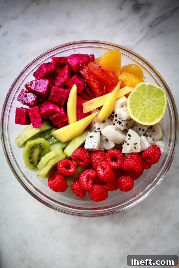 A vibrant and colorful tropical fruit salad elegantly presented in a clear glass bowl. This delightful medley features succulent red dragon fruit, sweet yellow dragon fruit, juicy mango, tangy kiwi, plump raspberries, and bright oranges.