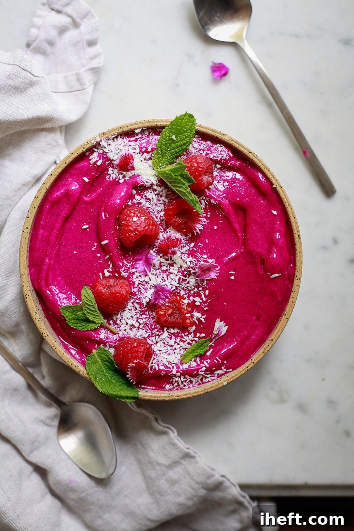 Beautiful photo of a bright pink pitaya smoothie bowl generously topped with fresh raspberries, crunchy granola, and delicate coconut shreds.