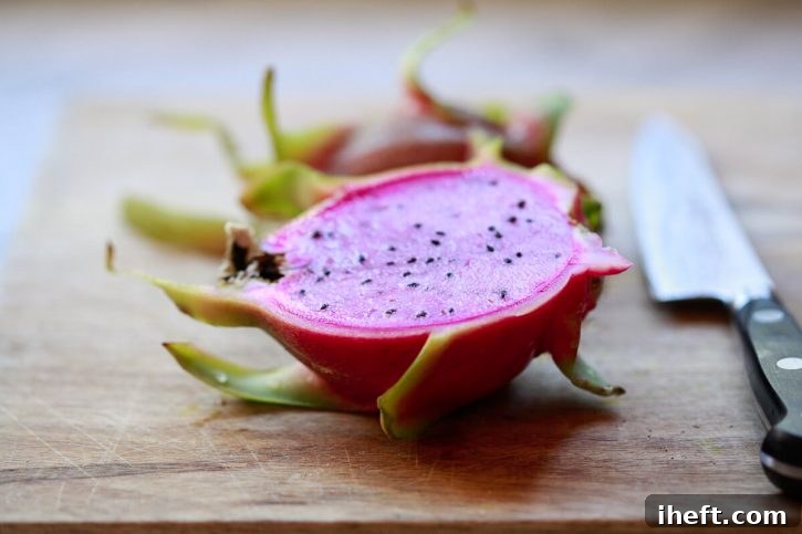 A dragon fruit with vivid pink flesh cut cleanly in half lengthwise, showcasing its delicate seeds on a rustic wooden cutting board.