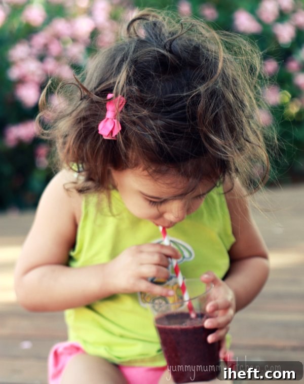 A child happily drinking a berry spinach smoothie from a glass with a straw, showing it's taste tester approved.