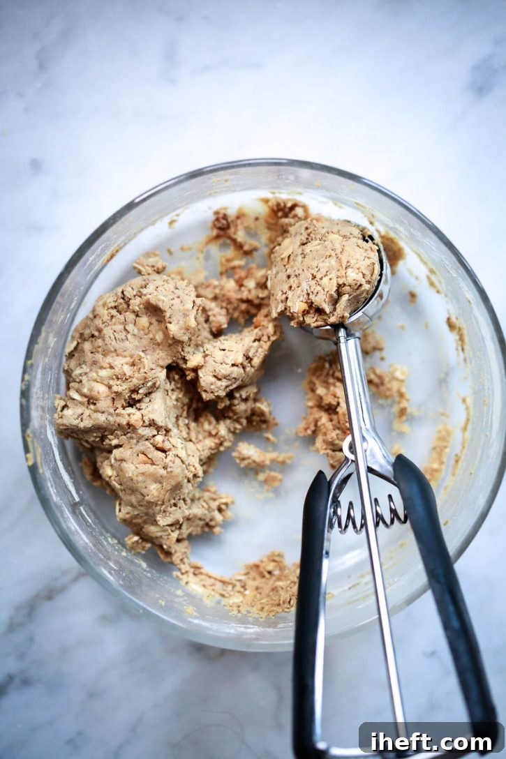 A small cookie scoop is perfectly poised, portioning out a ball of peanut butter oatmeal protein ball dough from a large mixing bowl. The hands in the image demonstrate the easy and efficient method of creating uniform protein balls for a healthy, homemade snack.