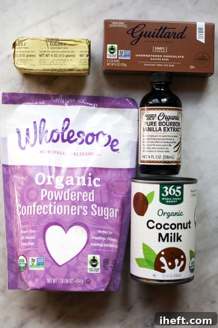 A pristine marble countertop displays the essential ingredients for vegan chocolate frosting: powdered sugar, solid blocks of vegan butter, a can of full-fat coconut milk, a bottle of vanilla extract, and squares of unsweetened chocolate. 