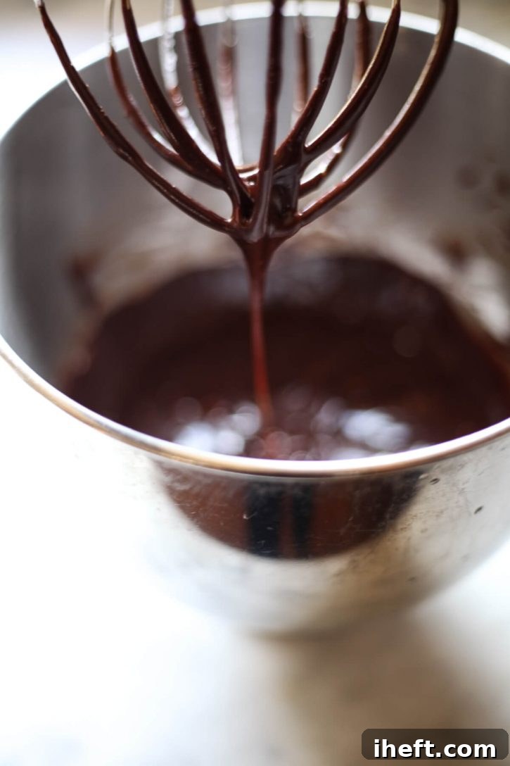 Homemade vegan chocolate frosting drips elegantly off a KitchenAid whisk attachment into a stainless steel mixing bowl, highlighting its smooth and luxurious consistency. 