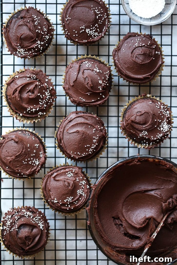 A collection of perfectly frosted vegan chocolate cupcakes are arranged on a metal cooling rack, with a bowl of the rich frosting placed casually to the side. 