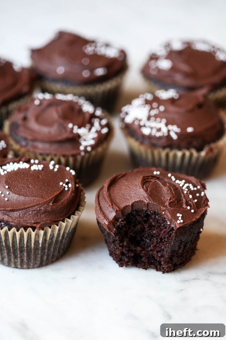 Photograph of vegan chocolate cupcakes with chocolate frosting and white sprinkles on a white counter. One cupcake has a bite out of it to show the inside. 