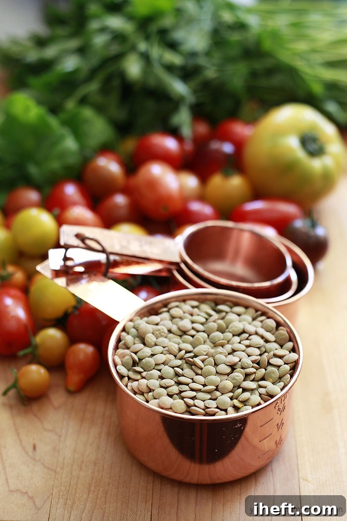 A copper measuring cup filled with green lentils next to tomatoes and parsley on a cutting board. 