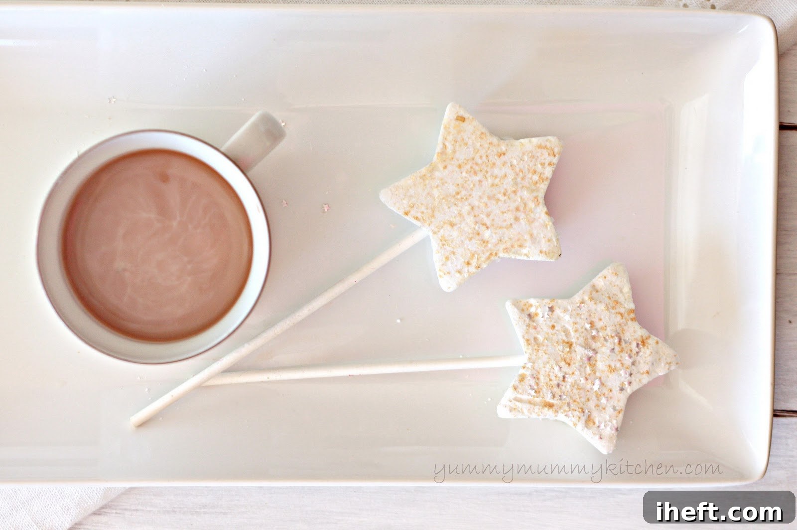 Two star-shaped marshmallows topped with edible gold glitter with sticks for stirring on a white platter next to a mug of hot chocolate.