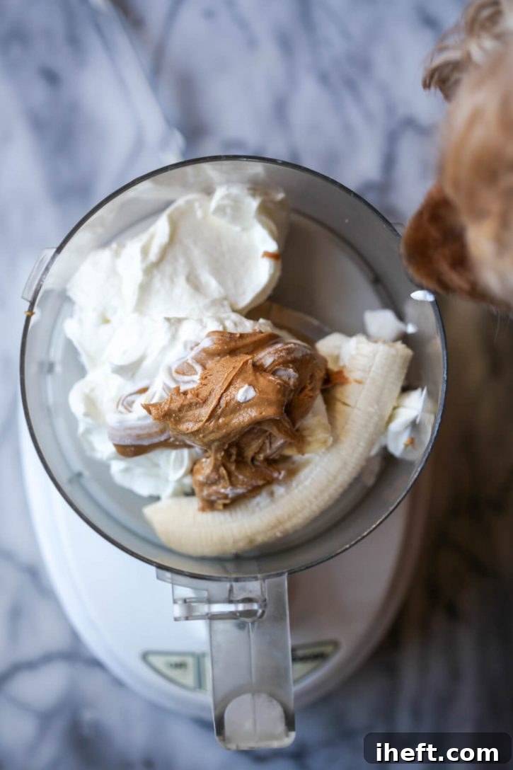 Greek yogurt, natural peanut butter, and a ripe banana are combined in a food processor, ready to be blended into homemade Frosty Paws dog ice cream. A curious dog peeks into the kitchen from the side.