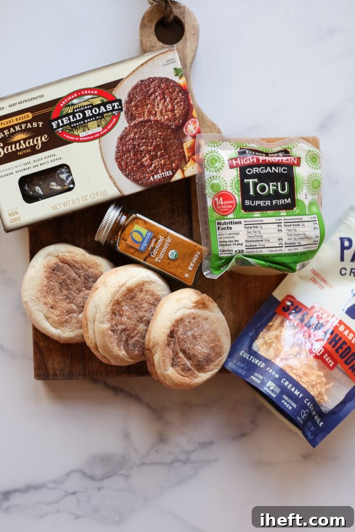 The ingredients for vegan breakfast sandwiches on a cutting board: Field Roast vegan breakfast sausage patties, high protein tofu, English muffins, turmeric, and Parmela's vegan cheese. 