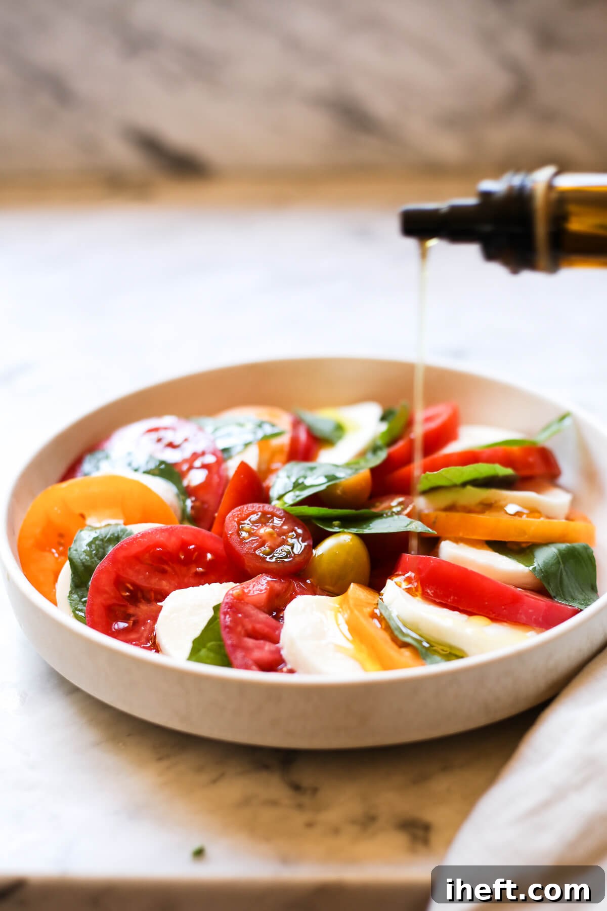 Olive oil is drizzled over a freshly made Caprese salad on a white plate in a white kitchen.
