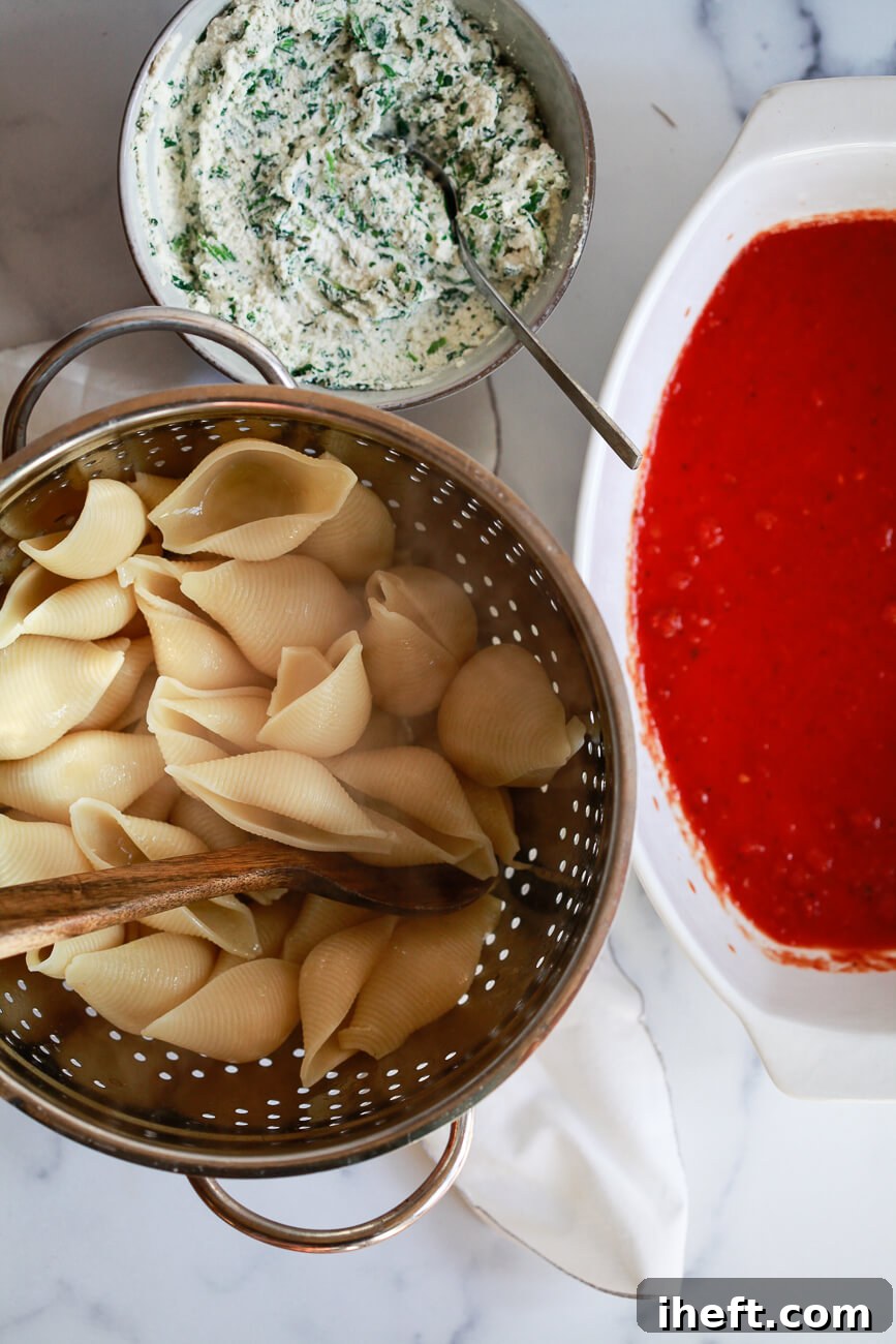 An overhead photo of cooked shell pasta in a colander, a bowl of ricotta filling, and a casserole dish filled with marinara sauce. 
