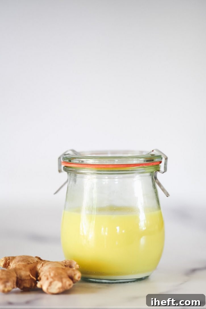 A small Weck jar filled with fresh, pure ginger juice sits elegantly on a marble counter, accompanied by a piece of fresh, unjuiced ginger root, highlighting the natural source of the potent elixir.