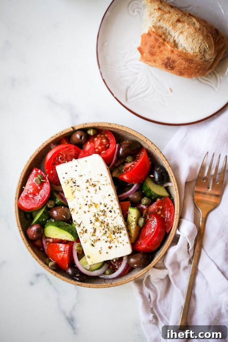 An inviting overhead photo of a freshly prepared traditional Greek salad (Horiatiki) generously topped with a large slab of feta cheese and sprinkled with dried oregano, elegantly presented on a marble countertop alongside a piece of rustic baguette.