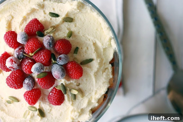 A vibrant top-down view of a festive Thanksgiving trifle, lavishly garnished to perfection. It features bright red fresh raspberries, glistening sugared cranberries, and crunchy green pepitas (pumpkin seeds), creating a rich tapestry of autumn colors and enticing textures, ready to be served.
