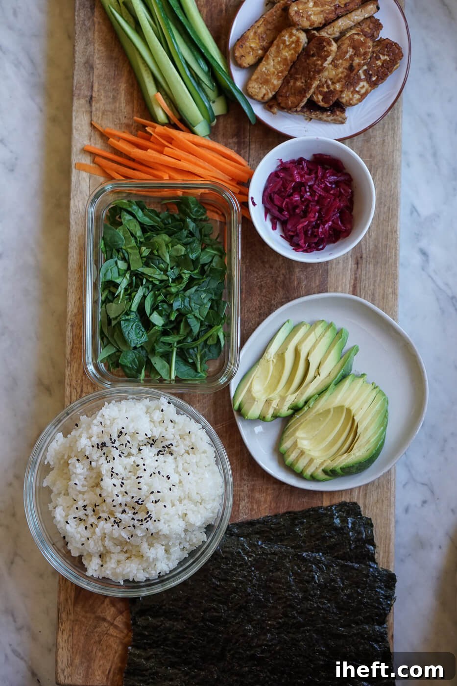 The ingredients for homemade sushi burritos ready on a cutting board: nori, rice, avocado, spinach, carrots, cucumber, red cabbage kraut, and tempeh.