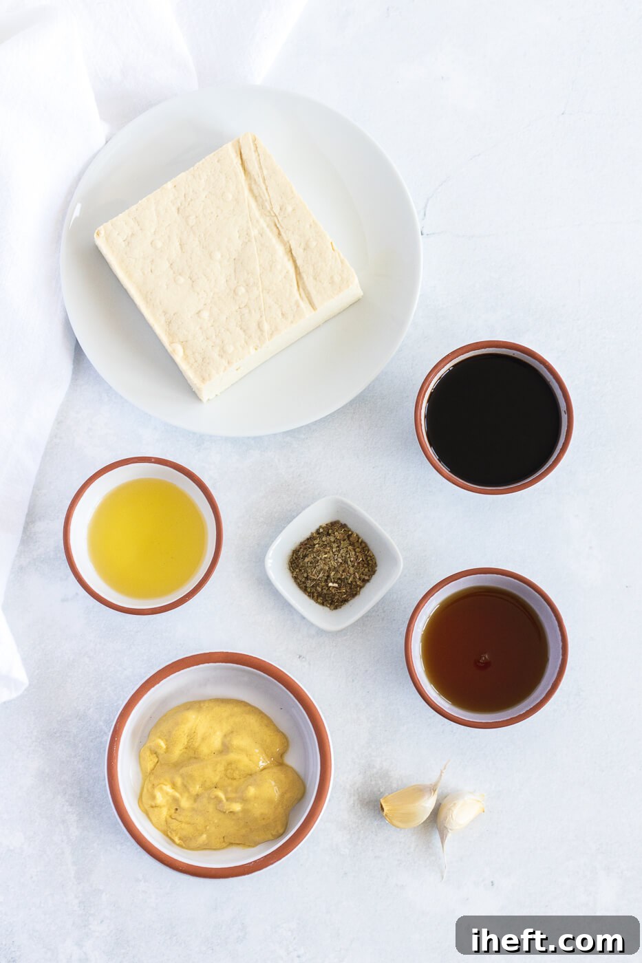 The ingredients for marinated tofu are pictured in small bowls on a white countertop next to a block of tofu. 