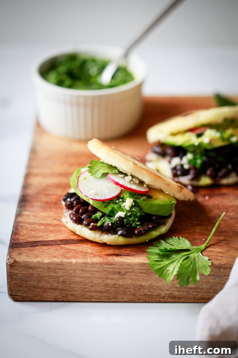 Two golden-brown Arepas Venezolanas generously filled with savory black beans, creamy avocado slices, a vibrant chimichurri sauce, crumbled queso fresco, and delicate radish slices, presented on a rustic wooden board.