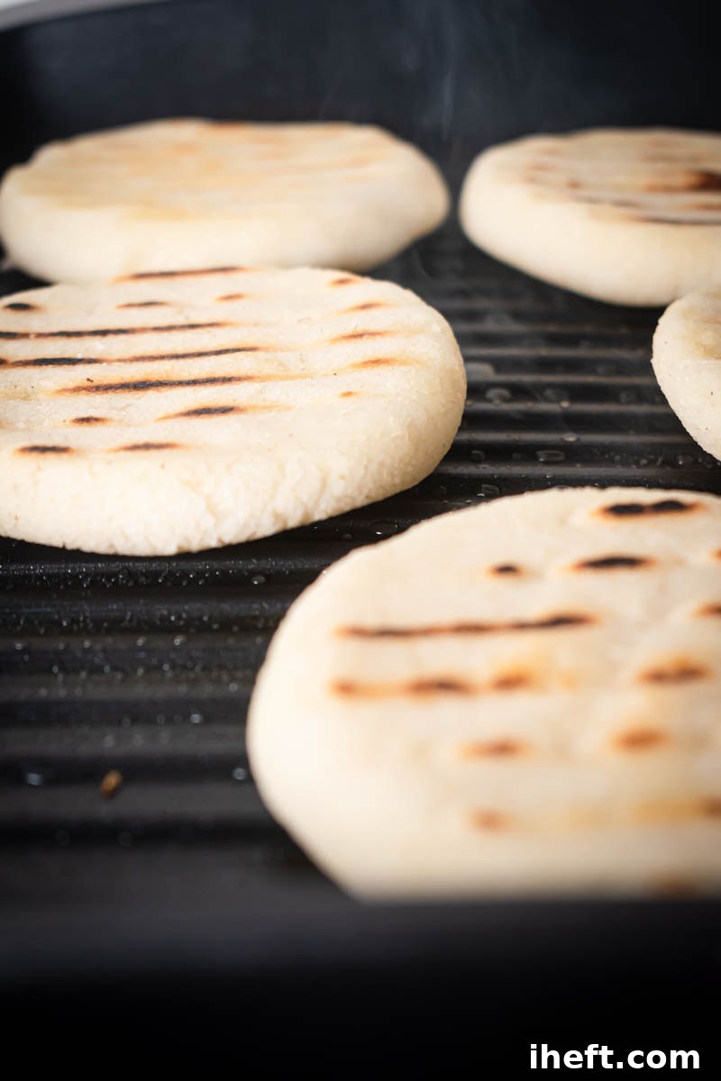 Several Arepas Venezolanas are sizzling and grilling on a hot, oiled grill pan, achieving beautiful golden-brown marks and a perfectly crispy texture.