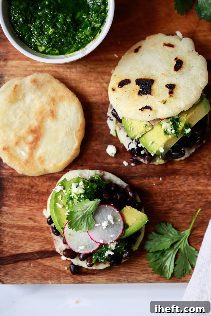 A selection of homemade Arepas Venezolanas, generously topped with savory black beans and creamy avocado, arranged on a cutting board beside a small bowl of vibrant green chimichurri sauce, showcasing a popular and delicious filling combination.