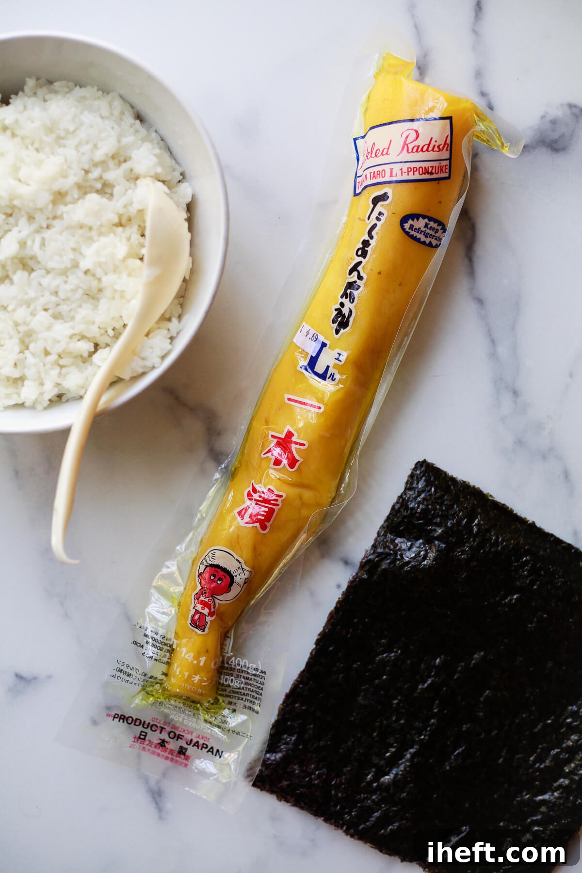 A bowl of perfectly cooked sushi rice, a package of vibrant oshinko, and sheets of nori seaweed sit on a sleek marble countertop, ready for sushi making.
