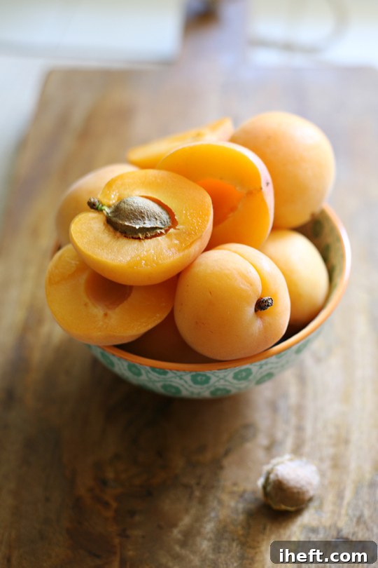 A small bowl filled with apricots sits on a wooden cutting board. One apricot is cut in half. 