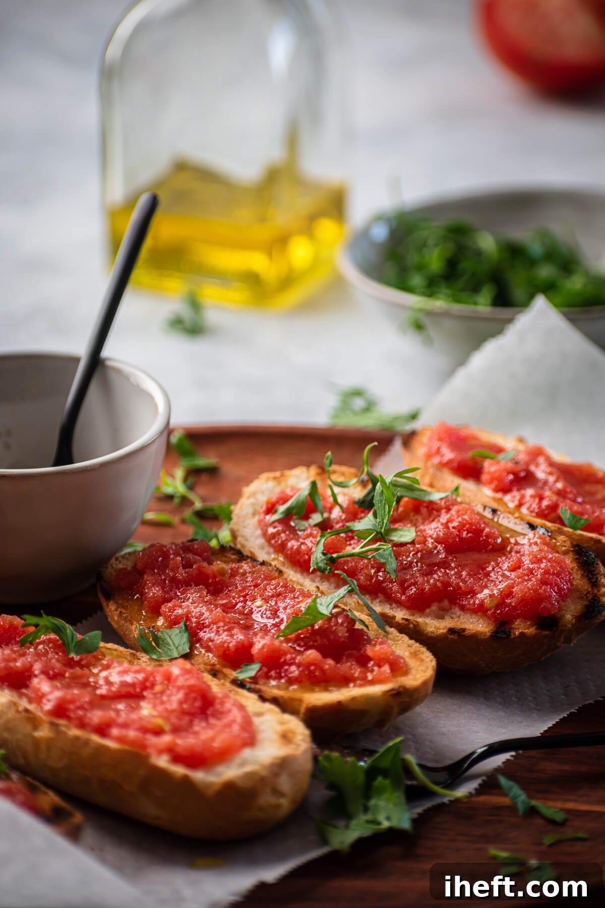 A beautiful food photo of four slices of pan con tomate garnished with fresh parsley. A jug of olive oil in the background. 