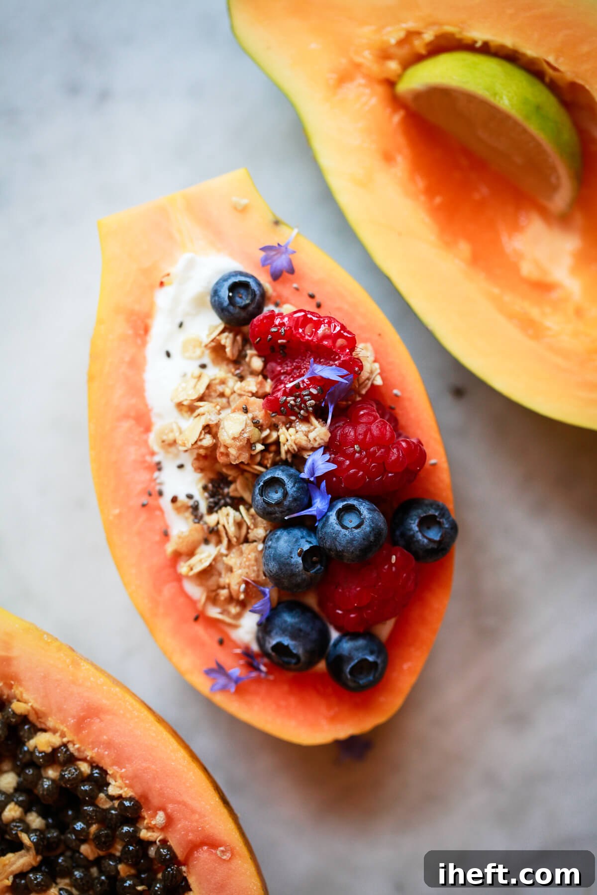 Three papaya halves artfully arranged on a marble countertop. One is garnished with fresh lime, another is a vibrant papaya boat filled with yogurt and granola, and the third showcases its intriguing seeds.