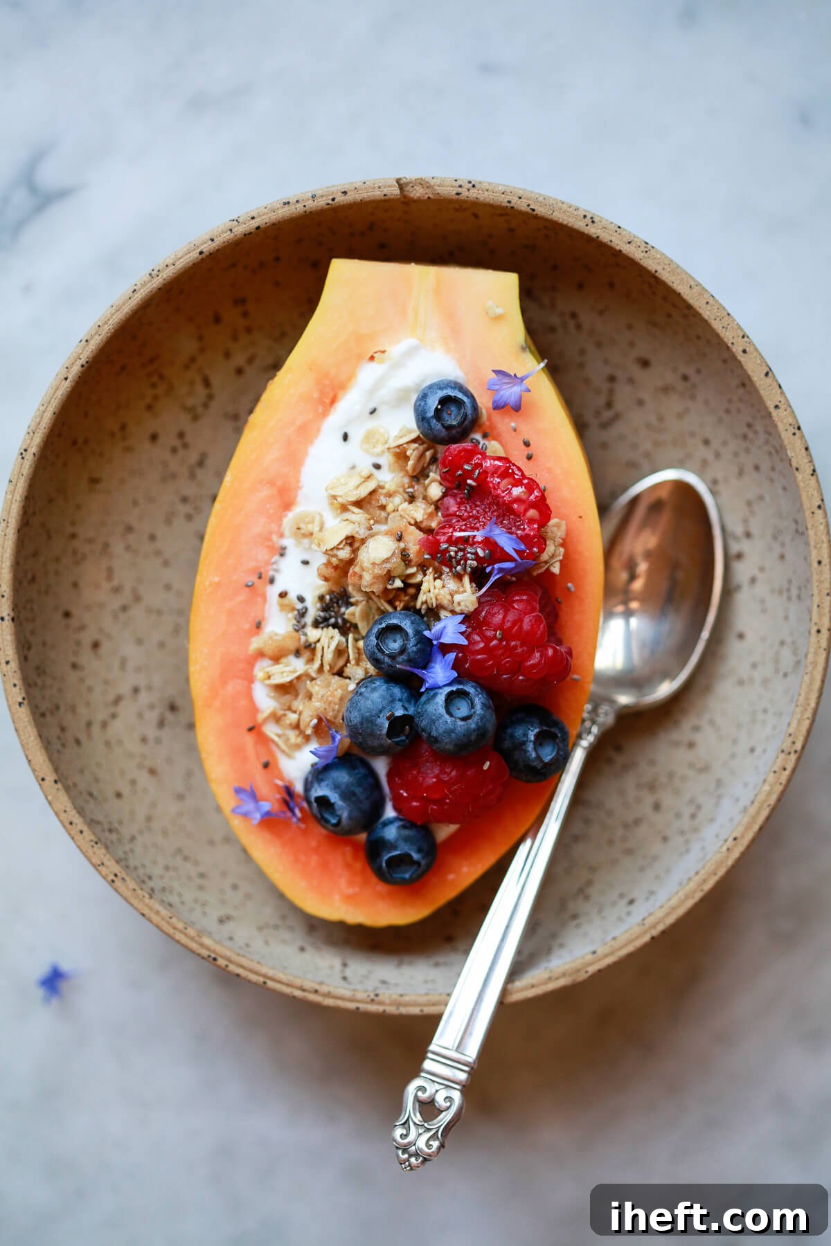 A beautifully prepared papaya boat, brimming with creamy yogurt, crunchy granola, and vibrant berries, nestled in a stone bowl with a silver spoon.