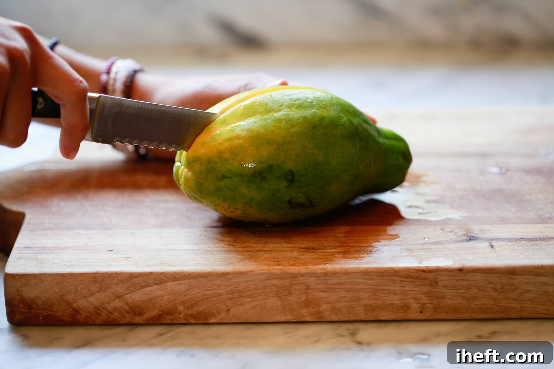 A sharp knife carefully cuts a vibrant green and yellow papaya in half on a wooden cutting board.
