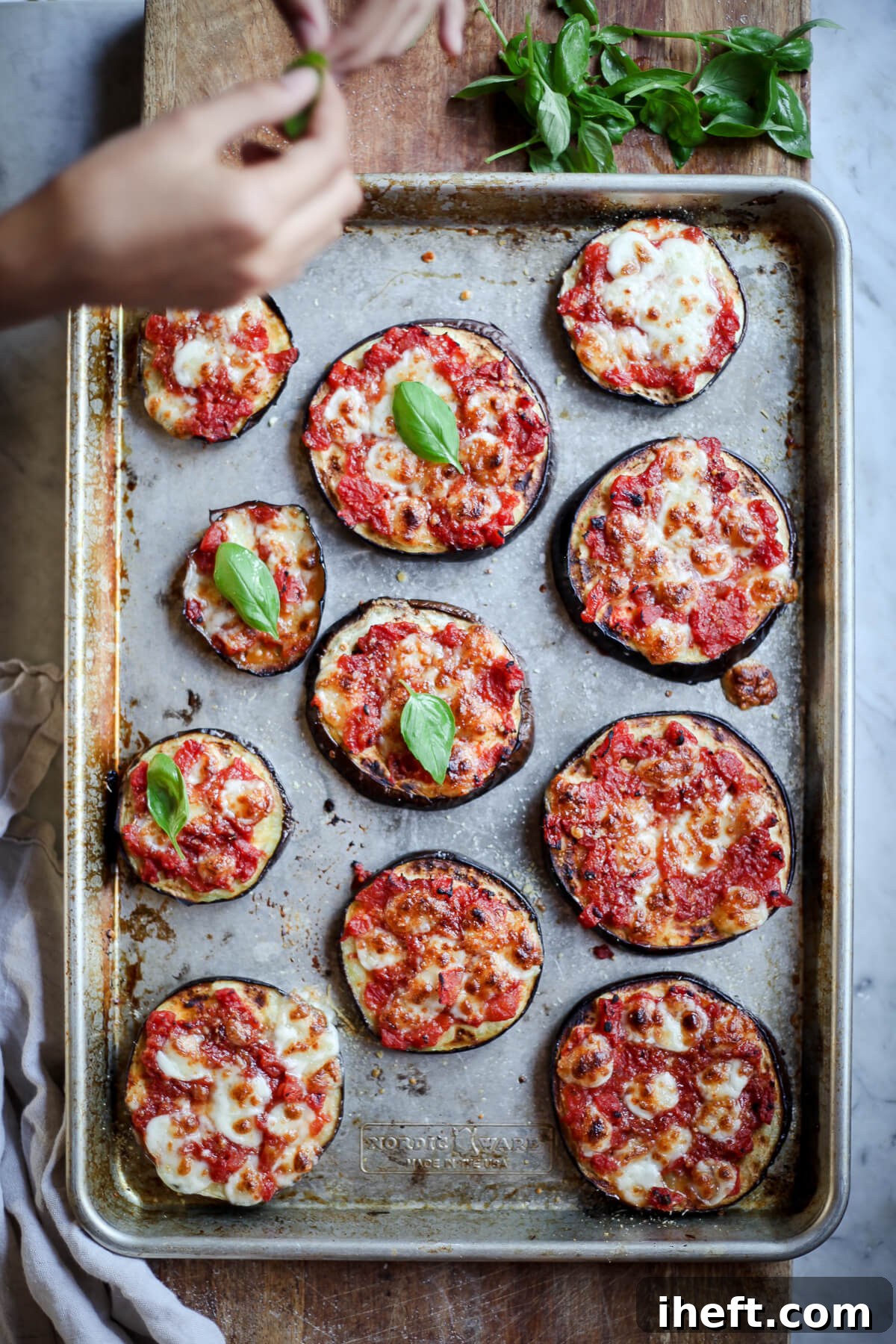 An overhead photo of Julia Child's eggplant pizza bites on a baking sheet while hands top them with fresh basil. 