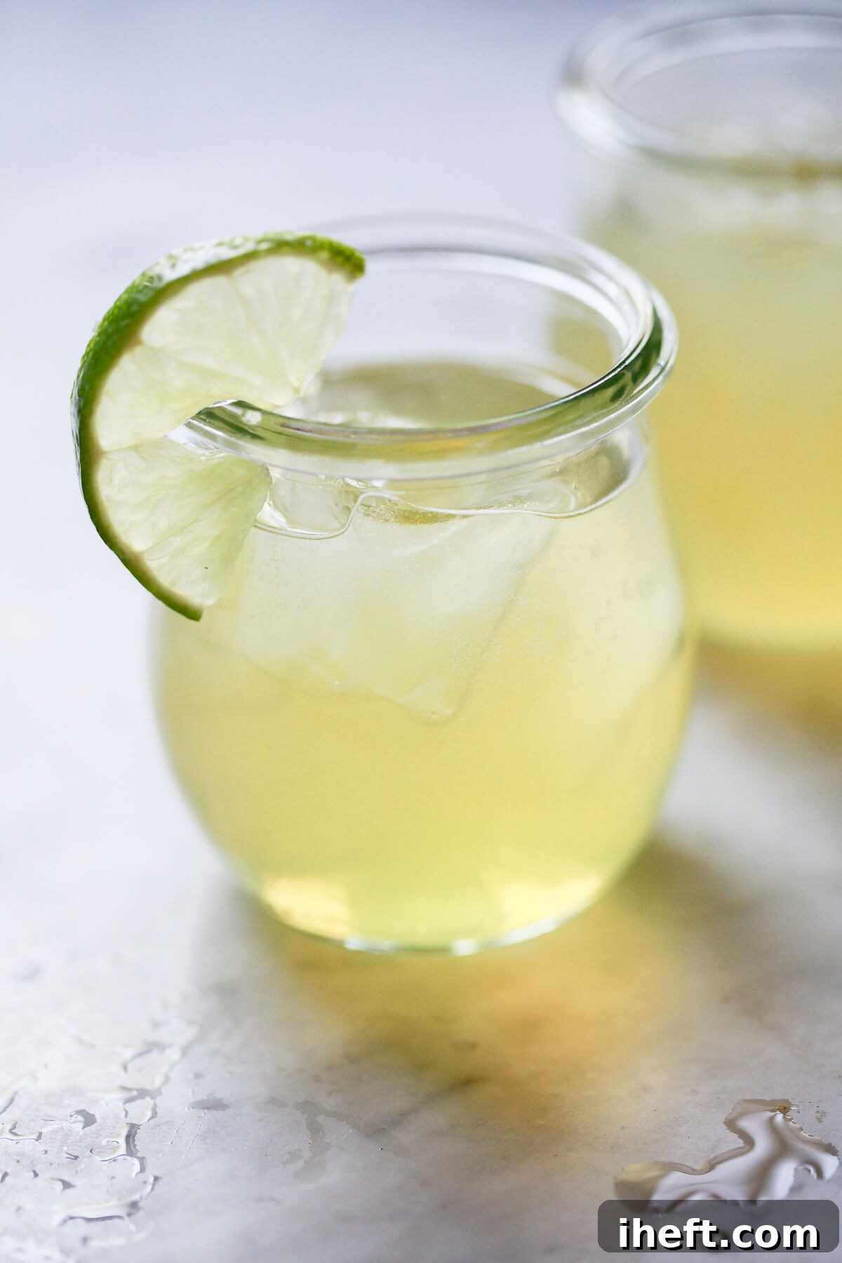 A freshly made green tea shot recipe in two small shot glasses garnished with lime sit on a marble countertop. 