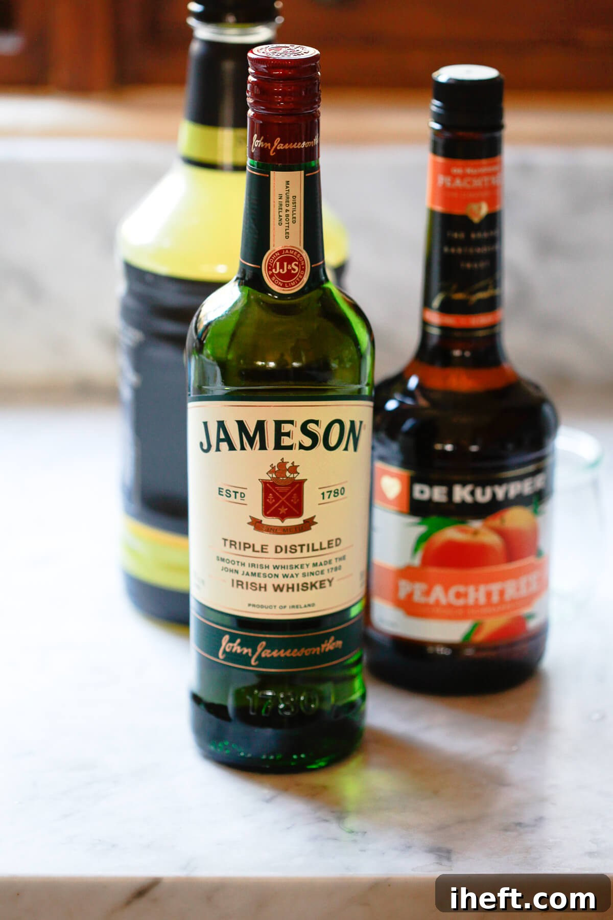 Three alcohol bottles sit on a marble countertop: Jameson Irish Whiskey in the foreground and peach schnapps and sweet and sour in the background. 