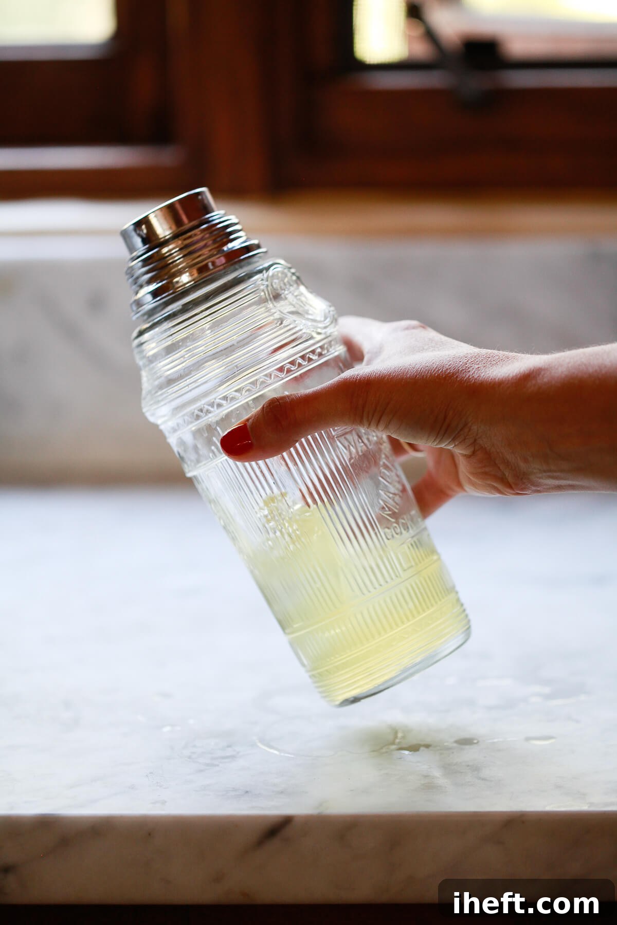 A hand shakes a vintage crystal cocktail shaker with ice and a yellow green tea shot in a white kitchen. 
