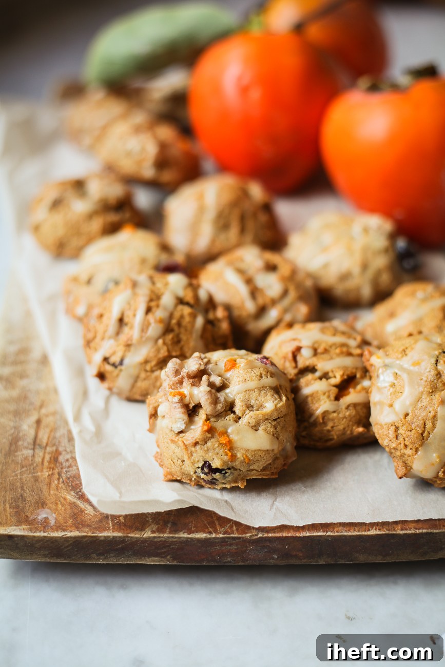 Soft and moist persimmon cookies with walnuts and cranberries, drizzled with a bright orange glaze, beautifully arranged on a rustic wooden cutting board with fresh Hachiya persimmons in the background. Ready for a delightful fall snack.