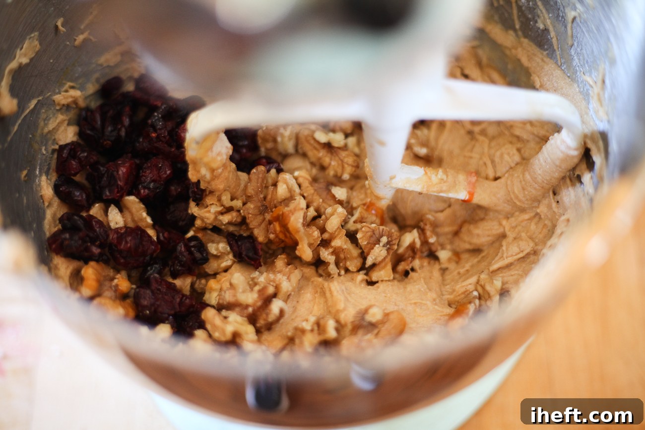 Chopped walnuts and vibrant dried cranberries are folded into the light orange persimmon cookie dough in a mixing bowl, adding texture and flavor.