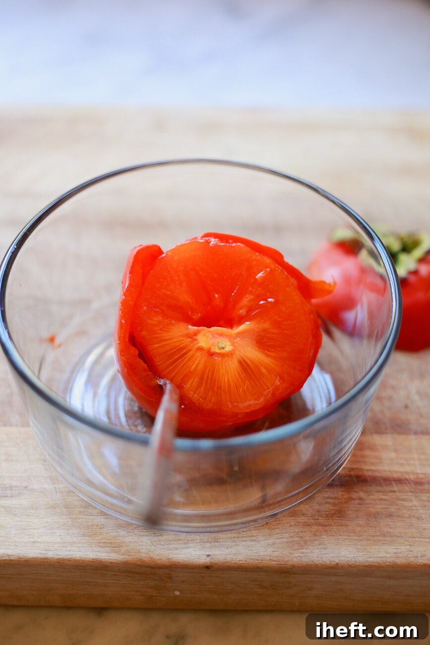 A spoon gently scoops the soft, translucent, and jelly-like flesh of a perfectly ripe Hachiya persimmon directly from its skin into a clear glass bowl, illustrating the ideal consistency for baking.